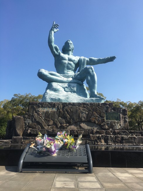 Bronze statue in the Nagasaki Peace Park of a human being sitting on a rock with one arm straight out and the other raised towards the sky. Boquets of flower sit below.